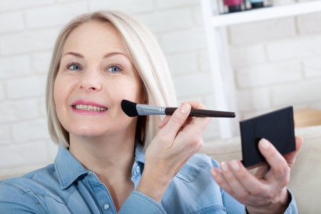 Feminine Grace. A mesmerizing photo capturing a woman's delicate hands expertly adorning her face with makeup, as she prepares for a special occasion. Woman is doing makeup in front of the mirrorの写真素材