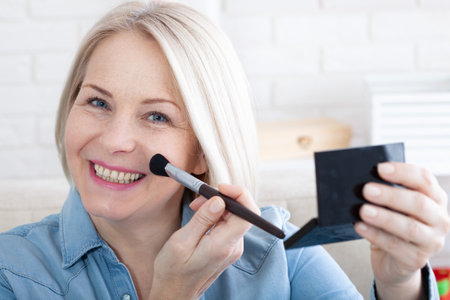 Beautiful happy woman engrossed in her makeup routine, with the mirror reflecting her poised and confident aura. Woman is doing makeup in front of the mirror at homeの写真素材
