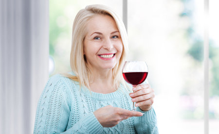 Happy woman holds a glass of red wine and looks relaxed at the camera sitting near the window.の写真素材
