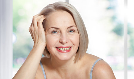 Happy beautiful middle-aged blonde woman shows off her perfectly well-groomed face and sitting relaxed near the window. Realistic images.の写真素材