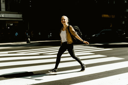 Young woman crossing a street in New York City, wearing a jacket and black pants.の素材