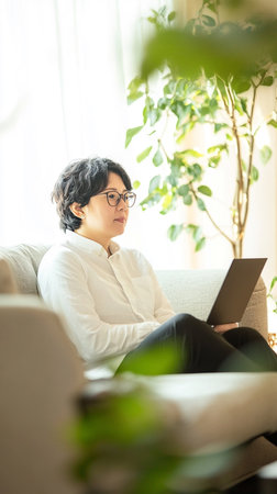 Asian woman using laptop while sitting on sofa in living room at homeの素材