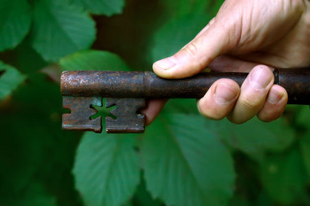 Human hand holding old rusty key, closeup shot with foliage blurred background