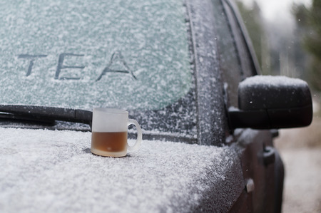 Cup of hot tea on a snowy car hood, concept of engine start in cold weatherの写真素材