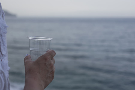 Plastic glass of water in female hand sea in the blurred background selective focus shotの写真素材