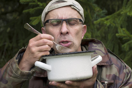 Matured man in camouflage and cap eating from casserole in the forest, outdoor horizontal shotの写真素材
