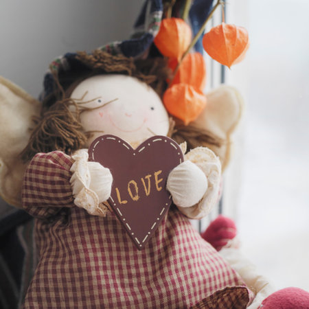 Doll sitting next to window and holding heart shape with word Love in hands, indoor square shot