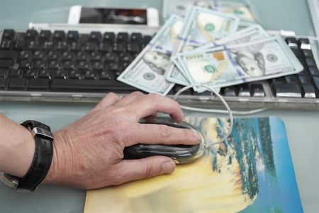 Male hand with a computer mouse next to a computer keyboard and US currency spread out on a top of it. Studio particular focus shot, concept of working online for moneyの写真素材