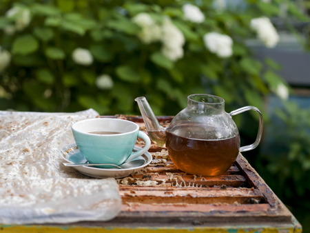 An open beehive with tea pot and tea cup with saucer on top of it, flowering garden in the blurred backgroundの写真素材