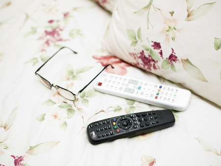Remote controls and eye wear laid on the bed linen, closeup with shallow depth of fieldの写真素材