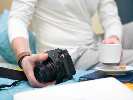 Man sitting on the bed or sofa with a photo camera in one hand and a cup of espresso in other,...