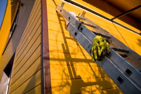 Yellow rubber gloves laid on the telescopic ladder next to the fresh painted wall of a wooden house
