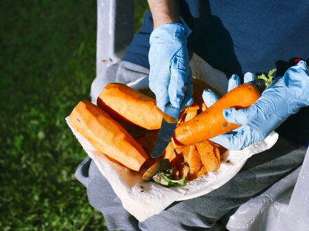 Female hands in protective gloves peeling carrot, outdoor close-upの写真素材