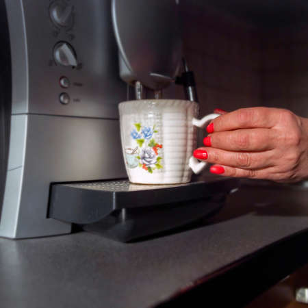 Female hand with manicured nails holding a mug next to a coffeemaker, indoor closeup