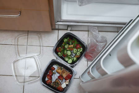 Food in plastic containers seemingly gone off on a floor next to open fridge