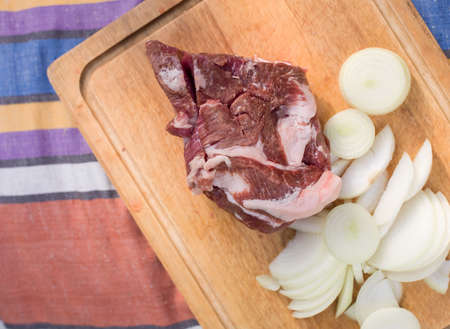 A piece of pork and sliced onion on a wooden chopping board, closeup still