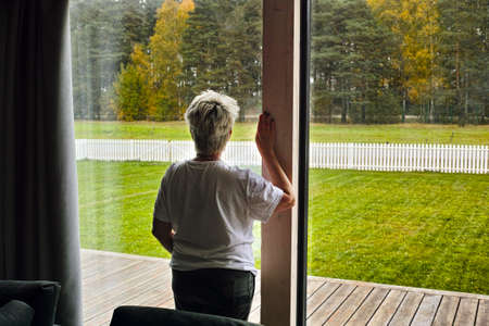 a woman standing near a window and looking at trees turning on yellow, indoor framed shotの写真素材