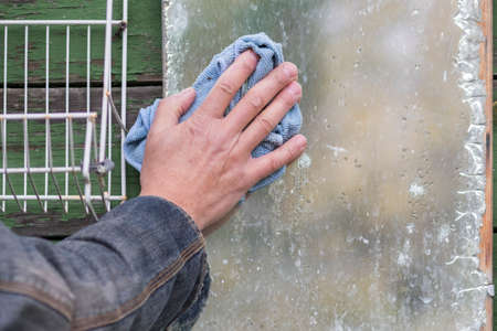 A male hand with rag wiping old damaged mirror, outdoor close upの写真素材