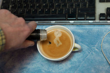 close up of a hand adding alcohol in a coffee cup placed near computer keyboardの写真素材