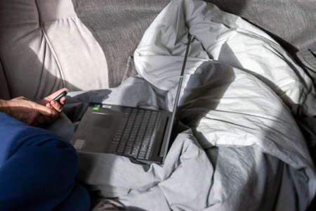 a man sitting on a messy unmade bed and working on a laptopの写真素材