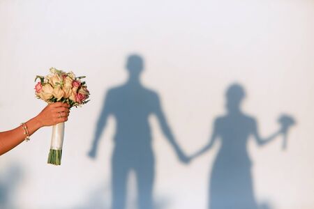 Hand with a bracelet holds a wedding bouquet. Shadows of a man and woman holding hands on a white wall background.の写真素材