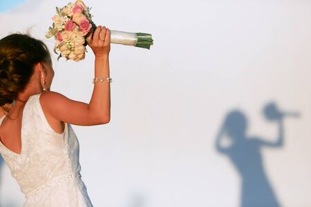 A dark-haired bride with a bouquet in her hand stands in front of a white wall with her shadow.の写真素材