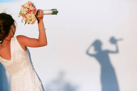 A dark-haired bride with a bouquet in her hand stands in front of a white wall with her shadow.の写真素材