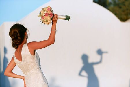 A dark-haired bride with a bouquet in her hand stands in front of a white wall with her shadow.の写真素材