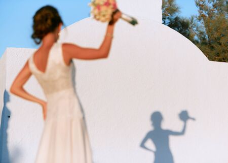 A dark-haired bride with a bouquet in her hand stands in front of a white wall with her shadow.の写真素材