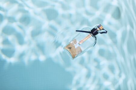 Sand and a note are inside the bottle, which is closed by a cork and floats in blue water.の写真素材