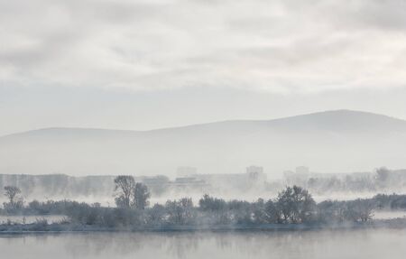 Winter landscape with foggy rivers is in the background of islands with trees, buildings and hills.の写真素材