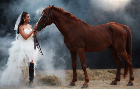 Young beautiful beauty bride in fashion white wedding costume stand with handsome horse on black backgroundの写真素材