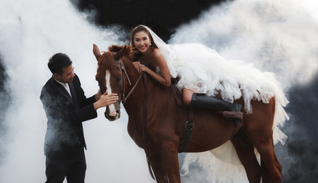Young newlywed couple, beautiful beauty bride in fashion white bridal wedding costume riding on strong muscular horse standing by smart and handsome groom on black dreamy backgroundの写真素材
