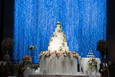 Wedding Cake on the table decorated by beautiful flowers at the stage with beautiful glittering blue light bokeh backdrop in wedding night ceremony. Wedding Ceremony tools and decorations concept.の写真素材