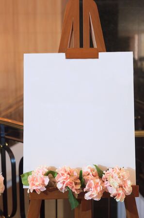 White blank empty canvas frame with flower decoration on wooden stand background. Use for Invitation Board Design in Wedding Reception Ceremony, Celebration Party. Creative Interior Designer concept.の写真素材