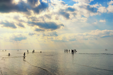 Silhouettes of people walking in the sea after the tide in the beautiful sunshine through the cloudsの写真素材