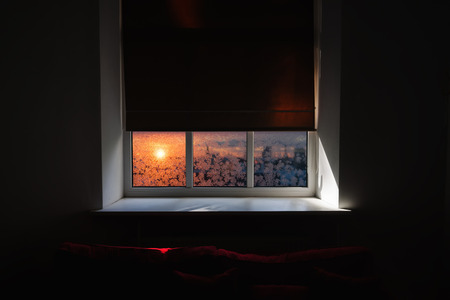 The window in the room at dusk with a red sofa near the window with a winter landscape through the frost on the streetの写真素材