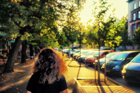 The girl with beautiful curly hair walking along the alley in backlit golden light through the green leaves of the trees next to the parked cars. Blurryの写真素材