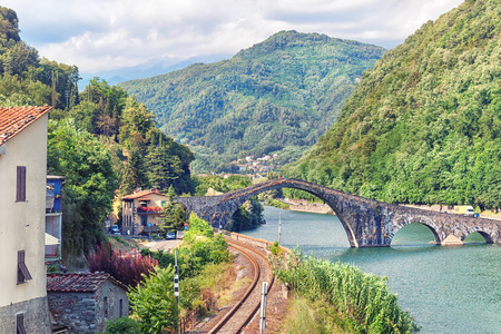 The railway past the houses along the river and the stone bridge of the Devil in the hills of Italy in a Sunny summer dayの写真素材