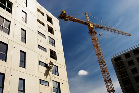 The worker is hanging on the wall of a multi-apartment building under construction against the background of a blue sky and a large craneの写真素材