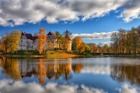 An ancient castle with towers on an island by the lake among autumn yellow trees with reflection in...