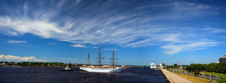 Panorama of the port of Riga on the Daugava river with a three-masted sailboat and a passenger ferry amidst beautiful clouds on a blue summer sky...の写真素材