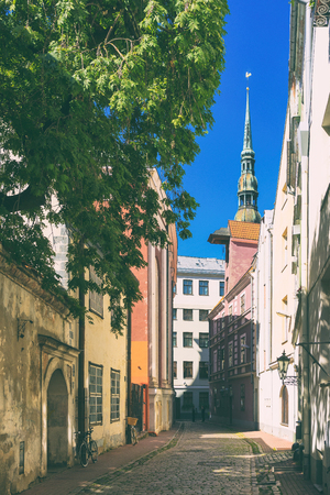 Street with old houses in the center of Riga in the summer against the backdrop of the Peter Church spire and green treeの写真素材