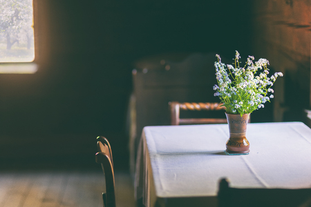 Wild flowers in a vase on a table with a white tablecloth in a simple wooden rural house on a sunny dayの写真素材