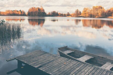 Wooden boat at the pier from gray boards on the lake with yellow trees on the shore in autumn. Latviaの写真素材