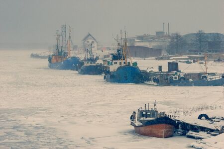Fishing boats stand in the ice of a frozen bay of the Baltic Sea on a background of snowfallの写真素材