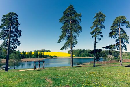 Wooden bridge on the blue water of a lake in spring. On the opposite bank is a yellow field of rapeseed. Blue horizon without clouds. Latviaの写真素材