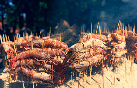 Shrimps are cooked on charcoal skewers at a street fair. Cooks and guests behind the smoke from the grill on the sand.の写真素材