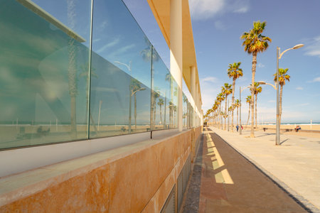 Promenade on the sandy beach of the Mediterranean in spring. The beach and vacationers are reflected in the glass facade of the building. Valenciaの写真素材