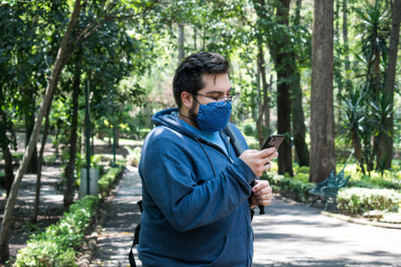 Front view of young Hispanic man walking down the street with handmade mask, checking application notifications on his cell phone.の写真素材